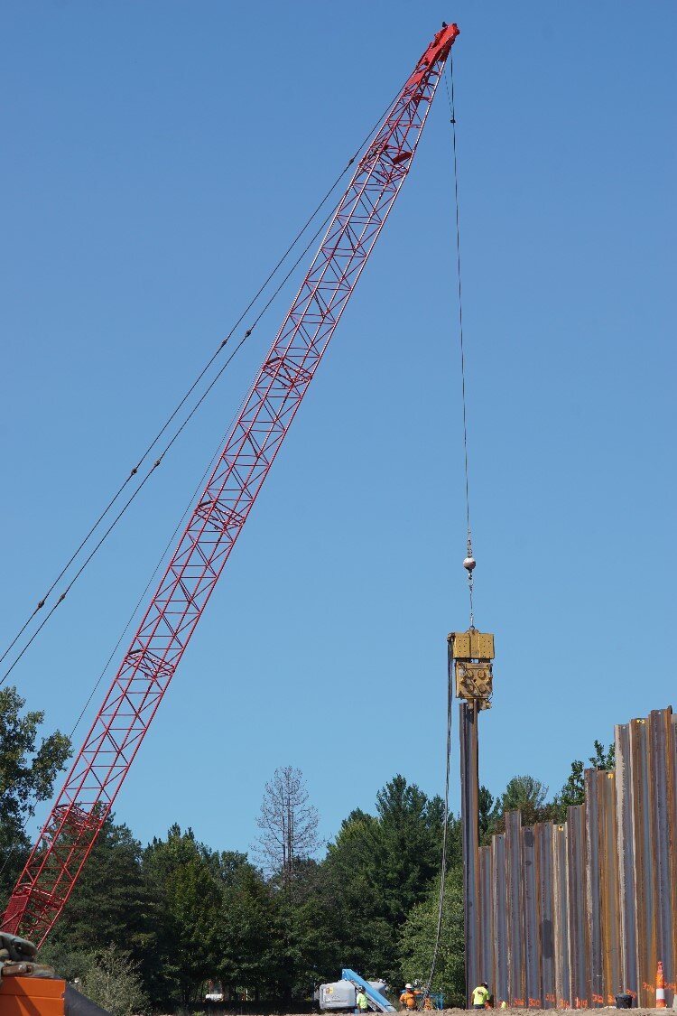 Steel sheet piling is drilled deep underground by a large crane. The sheet piling interlocks together to form an impermeable layer to protect against seepage in the instance of a future flood.