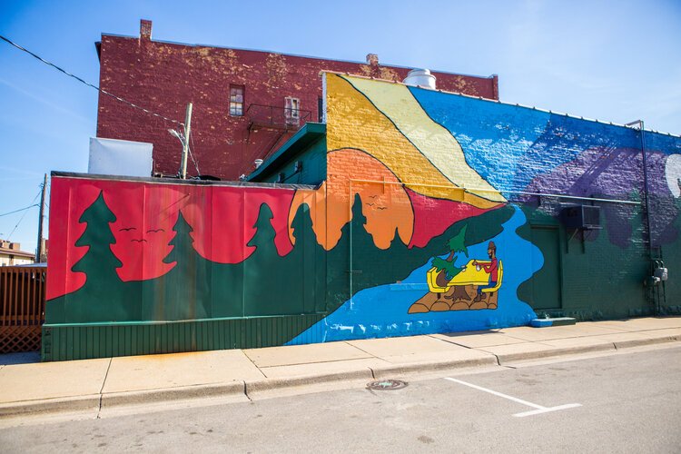 Along the west side of Zef's Coney Restaurant is a scene featuring a color-blocked sunset illuminating a man and a pine tree chatting. The mural honors Bay City's lumbering history.