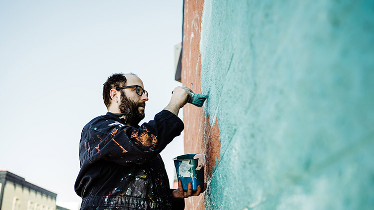 Artist Adam Wernecke painting on mural on the building housing Asian Noodle. Located on Saginaw Street in downtown Bay City, surrounding businesses offer sushi, art, coffee, craft beer, and artisan chocolates and cheeses.